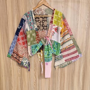 May include: A patchwork kimono-style top with a tie closure. The garment features a variety of colorful fabric patterns, including floral, geometric, and paisley designs. The sleeves are wide and the top is hanging on a wooden hanger.