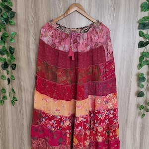 May include: A long, tiered skirt with a patchwork design. The skirt features various shades of red, pink, and orange, with floral and patterned fabric sections. It hangs from a wooden hanger against a light wood-grain background.