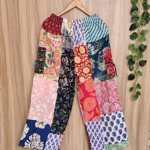 May include: Patchwork pants with a variety of floral and geometric patterns in shades of red, pink, blue, and green. The pants are hanging on a wooden hanger against a light brown wooden background.