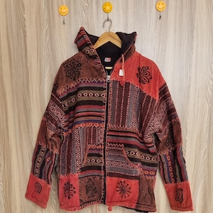 May include: A patchwork hooded jacket with a zipper closure. The jacket features a variety of patterns and colors, including red, brown, and black. It has a drawstring at the waist and pockets. The jacket is hanging on a wooden hanger.