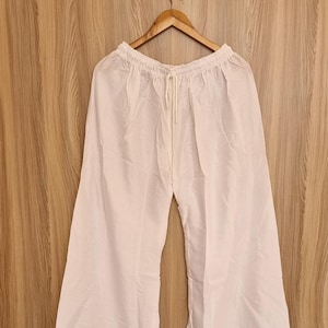 May include: White, wide-leg trousers with a drawstring waist hang on a wooden hanger. The trousers are made of a lightweight fabric and are displayed against a wood-grain background. A yellow smiley face hook is above the hanger.