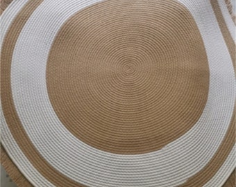 May include: A round, natural fiber rug with a white and beige striped border. The rug has a fringed edge.
