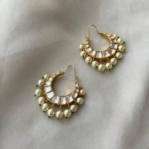 May include: A pair of gold-toned hoop earrings. Each earring features a crescent shape with clear, rectangular stones and dangling faux pearls. Small green beads accent the design. The earrings are displayed on a white, textured surface.