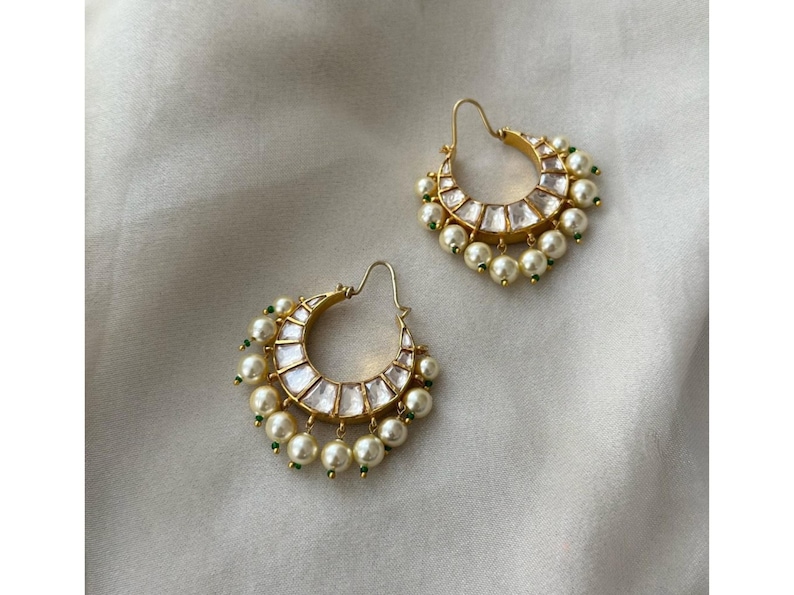 May include: A pair of gold-toned hoop earrings. Each earring features a crescent shape with clear, rectangular stones and dangling faux pearls. Small green beads accent the design. The earrings are displayed on a white, textured surface.