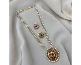 May include: A pearl necklace and earring set. The necklace has multiple strands of small, white pearls. The pendant and earrings have a gold-coloured setting with a central red stone, surrounded by smaller pearls.