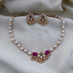 May include: A gold necklace with a pearl chain and a pendant featuring pink and green stones. Matching earrings with a similar design are included.