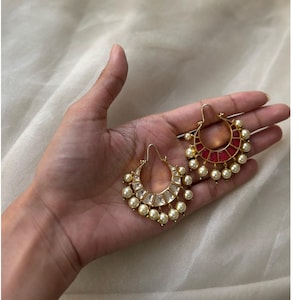 May include: A pair of gold-toned hoop earrings with pearl and gemstone accents. One earring features a red gemstone design, while the other has clear gemstones. The earrings are displayed on a hand against a soft, neutral background.