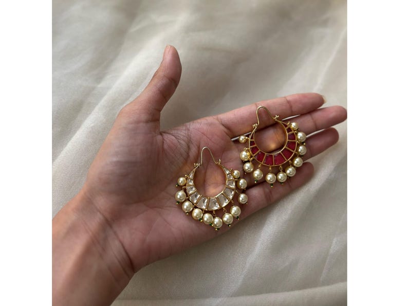 May include: A pair of gold-toned hoop earrings with pearl and gemstone accents. One earring features a red gemstone design, while the other has clear gemstones. The earrings are displayed on a hand against a soft, neutral background.