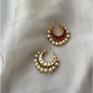 May include: Two gold-toned hoop earrings with a crescent shape. One earring features a red panel design, while the other has a white panel design. Both are adorned with small, round, white pearls and green accents.