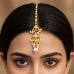 May include: A gold-toned headpiece with a central vertical chain and a decorative pendant. The pendant features clear, teardrop-shaped stones and small, round pearls. A large pearl hangs at the bottom. The headpiece is set against dark hair.