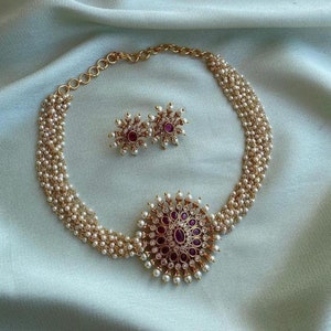 May include: A gold necklace with a multi-strand pearl design and a large, round pendant with a pink stone surrounded by white stones. Matching earrings with pink stones are also included.