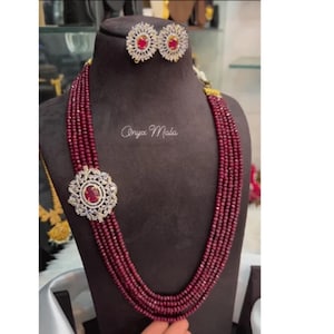 Puede incluir: Un collar de cuentas color granate con un gran colgante redondo que presenta una piedra roja rodeada de brillantes transparentes. El collar está formado por múltiples hileras de cuentas y tiene pendientes a juego con piedras rojas y brillantes transparentes.