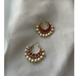 May include: A pair of gold-toned hoop earrings. Each earring features a crescent-shaped design with a red center and is adorned with white pearls and small green accents. The earrings are displayed on a white, textured surface.