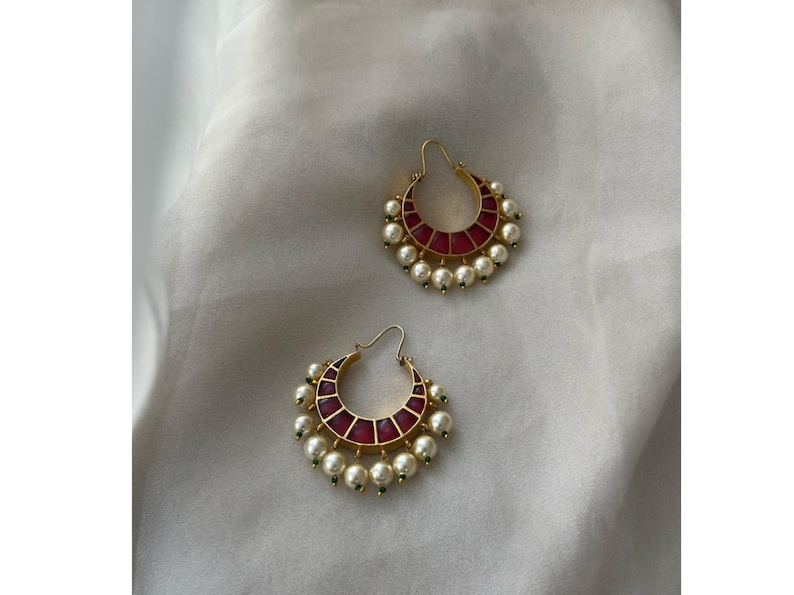 May include: A pair of gold-toned hoop earrings. Each earring features a crescent-shaped design with a red center and is adorned with white pearls and small green accents. The earrings are displayed on a white, textured surface.