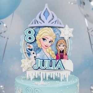 May include: A light blue birthday cake with a decorative topper featuring Elsa and Anna from Frozen, the number 8, and the name "Julia." The cake is decorated with snowflakes and swirls. Matching plates and napkins are also visible.