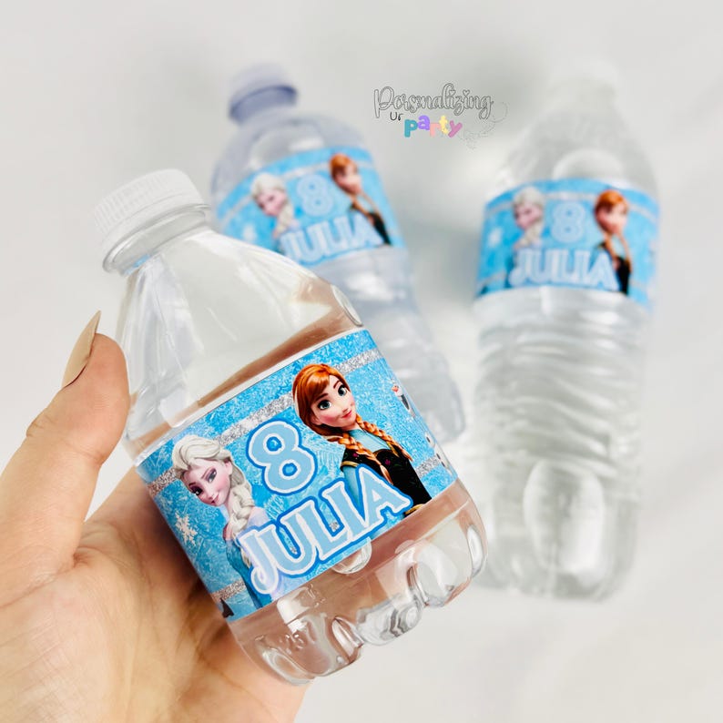 Peut inclure: Trois bouteilles d'eau en plastique transparent avec des &eacute;tiquettes bleues personnalis&eacute;es repr&eacute;sentant des personnages de dessin anim&eacute;, le chiffre 8 et le nom JULIA en blanc. Les &eacute;tiquettes ont une bordure argent&eacute;e scintillante.