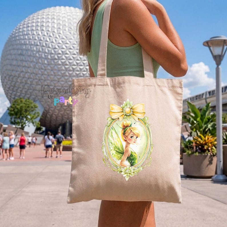 Peut inclure: Un sac fourre-tout en toile beige orn&eacute; d'un motif central repr&eacute;sentant une f&eacute;e aux cheveux blonds et aux ailes vertes, encadr&eacute;e dans un ovale d&eacute;coratif surmont&eacute; d'un n&oelig;ud jaune. Le motif est imprim&eacute; sur le devant du sac &agrave; deux anses.