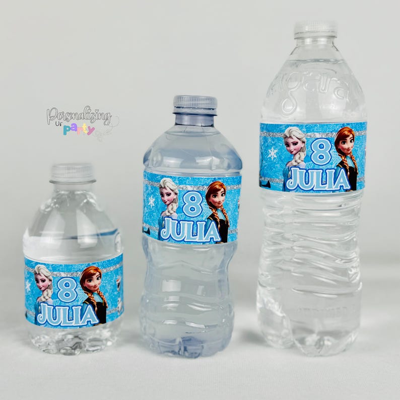 Peut inclure: Trois bouteilles d'eau en plastique transparent de tailles diff&eacute;rentes, chacune avec une &eacute;tiquette bleue &agrave; bordure scintillante, des flocons de neige et deux personnages de dessin anim&eacute;. Le num&eacute;ro 8 et le nom JULIA sont inscrits.