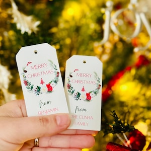 Może przedstawiać: Dwie białe etykiety prezentowe na Boże Narodzenie z kwiatowym wieńcem i napisem "Merry Christmas" w kolorze czerwonym i zielonym. Na jednej etykiecie napisano "from Benjamin", a na drugiej "from The Cunha Family". Etykiety są trzymane przed rozmytą choinką.