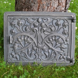 Puede incluir: Una puerta de estufa de hierro fundido con un intrincado diseño floral. La puerta es gris y tiene una bisagra decorativa.