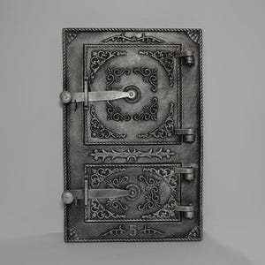 Op de afbeelding: Een zwarte gietijzeren ovendeur met sierlijke details en twee grendels. De deur heeft een "5" aan de onderkant.