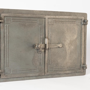 Peut inclure: Une porte métallique rectangulaire et patinée avec deux panneaux à charnières. La porte est de couleur grise sourde et présente des accents décoratifs rectangulaires et verticaux. Un loquet et une poignée fixent les portes, et des charnières sont visibles sur les côtés.