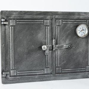 Puede incluir: Una puerta de horno de metal gris con un termómetro y un pestillo. La puerta tiene un diseño decorativo con líneas en relieve.