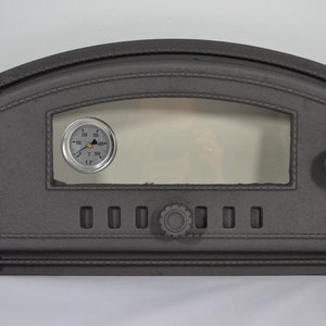 Op de afbeelding: Een zwarte gietijzeren pizza-ovent deur met een glazen raam en een ronde thermometer. De thermometer heeft een zwart gezicht met witte cijfers en een gouden naald. De deur heeft een knop aan de rechterkant en een reeks kleine gaten aan de onderkant.