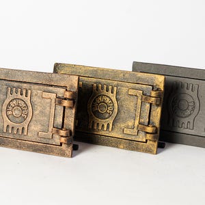 Op de afbeelding: Drie gietijzeren schouwdeuren in verschillende afwerkingen: antiek messing, goud en zwart. Elke deur heeft een decoratief ontwerp met een ronde opening en een vergrendeling.