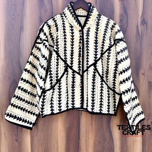 May include: Cream-colored jacket with a black geometric pattern of triangles and black trim. The jacket has a button-up front and long sleeves. The text "TEXTILES CRAFT" is visible on the lower right corner.
