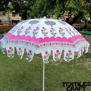 May include: A white and pink floral patterned outdoor umbrella. The umbrella features a scalloped edge with hanging floral accents. The design includes pink flowers and green leaves on a white background. The umbrella is supported by a white pole and is set on a grassy area.