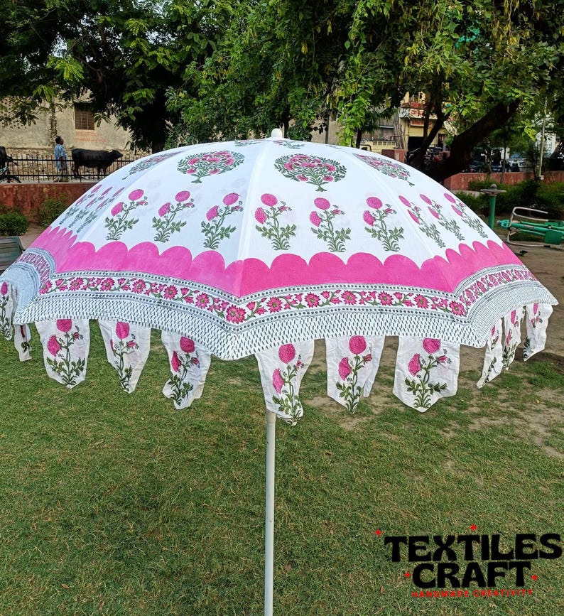 May include: A white and pink floral patterned outdoor umbrella. The umbrella features a scalloped edge with hanging floral accents. The design includes pink flowers and green leaves on a white background. The umbrella is supported by a white pole and is set on a grassy area.