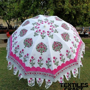 May include: A white outdoor umbrella with a pink floral pattern. The umbrella has a scalloped edge with a decorative border. The design includes pink flowers and green leaves. The umbrella is set up on a grassy area.
