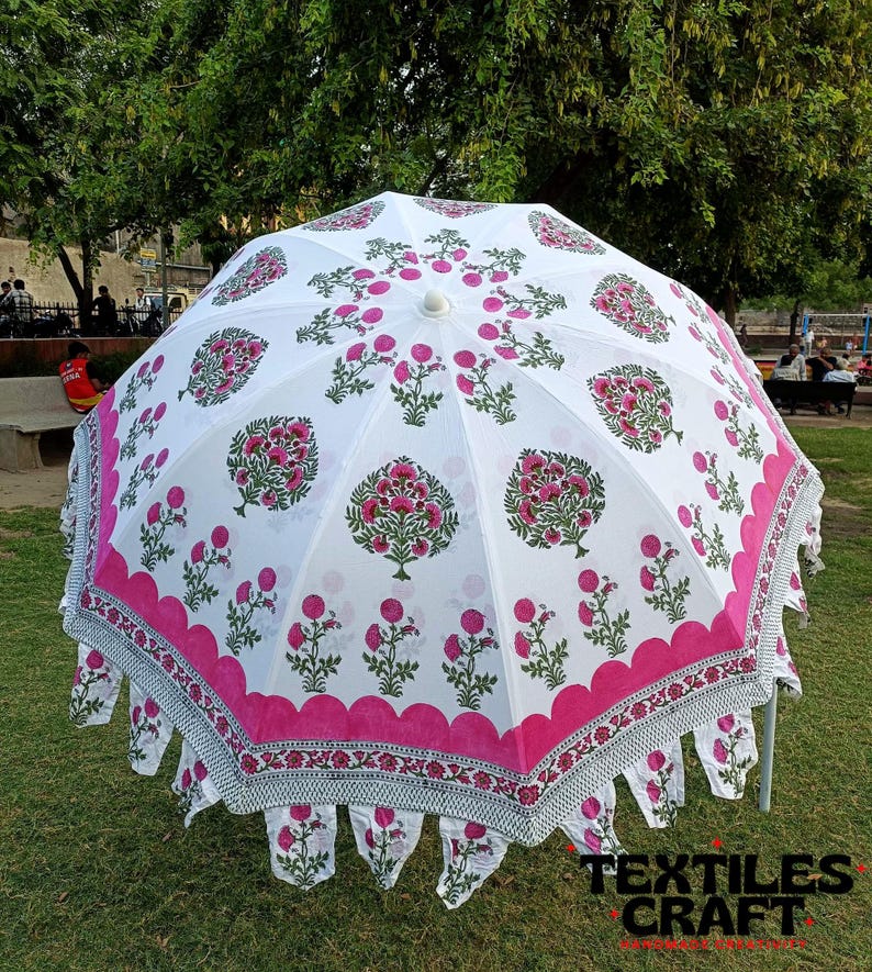 May include: A white outdoor umbrella with a pink floral pattern. The umbrella has a scalloped edge with a decorative border. The design includes pink flowers and green leaves. The umbrella is set up on a grassy area.