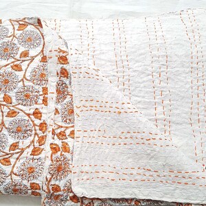 Puede incluir: Colcha kantha blanca con un patr&oacute;n floral en naranja y marr&oacute;n. La colcha est&aacute; cosida a mano con una puntada corrida en hilo naranja. La colcha est&aacute; doblada por la mitad y las puntadas son visibles en el lado posterior de la colcha.