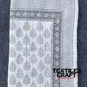 Op de afbeelding: Wit katoenen dekbed met een zwart en grijs bladmotief en een rand van geometrische ontwerpen. Het dekbed is dubbelgevouwen en heeft de tekst "TEXTILES + CRAFT + HANDMADE CREATIVITY +" gedrukt in de rechterbenedenhoek.