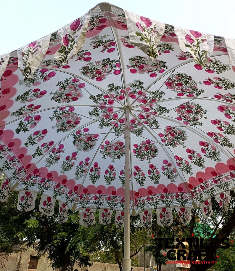 May include: A white umbrella with a floral pattern in pink and green. The umbrella has a scalloped edge with pink trim and decorative hanging fabric. The umbrella is open, showing the interior design.