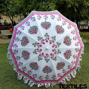 May include: A white umbrella with a floral pattern in pink and green. The umbrella has a scalloped pink border and a white handle. The text "TEXTILES CRAFT" is visible at the bottom right. The umbrella is open on a grassy surface.