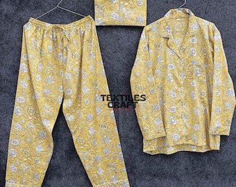 Op de afbeelding: Een gele pyjamaset met bloemenpatroon, bestaande uit een broek, een overhemd met knopen en een bijpassend zakje. De set is gemaakt van een zachte stof en wordt op een hanger gepresentteerd.