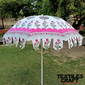 May include: A large, open white and pink floral-patterned umbrella. The umbrella features a scalloped pink border and decorative floral pendants. The design includes pink flowers and green leaves. The umbrella is set up on a grassy area.