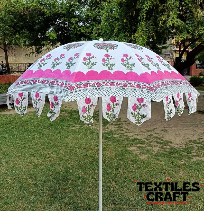 May include: A large, open white and pink floral-patterned umbrella. The umbrella features a scalloped pink border and decorative floral pendants. The design includes pink flowers and green leaves. The umbrella is set up on a grassy area.