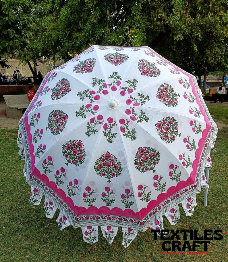 May include: A white outdoor umbrella with a floral pattern in pink and green. The umbrella has a scalloped edge with a pink border and decorative fringe. The umbrella is open and set up on a grassy area.
