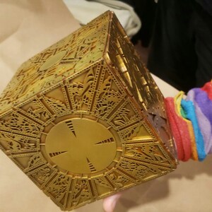 May include: A gold-colored metal puzzle box with intricate designs and a circular center. The box is being held by a hand wearing a rainbow striped sleeve.