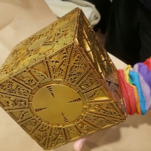 Könnte beinhalten: Eine goldfarbene Metall-Rätselbox mit komplizierten Designs und einem runden Zentrum. Die Box wird von einer Hand gehalten, die einen regenbogenfarbenen Ärmel trägt.