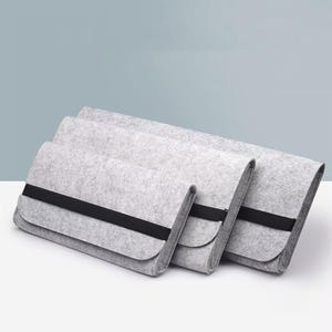 Peut inclure: Trois pochettes pour ordinateur portable en feutre gris avec une fermeture à bande élastique noire. Les pochettes sont disposées en rangée décalée, présentant leurs différentes tailles. Le feutre a un aspect doux et texturé.