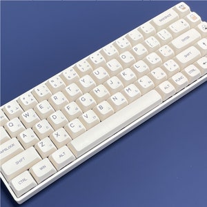 Puede incluir: Un teclado mecánico blanco con un juego de teclas de color crema. Las teclas tienen letras japonesas e inglesas. El teclado tiene una barra espaciadora blanca y un marco blanco.
