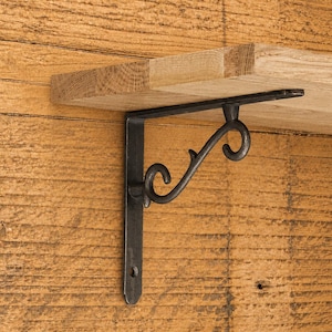 May include: A black metal shelf bracket with a curved, scroll-like support arm attached to a flat vertical mounting plate, holding a light-colored wooden shelf board. The bracket features a dark, matte finish and a simple, functional design.