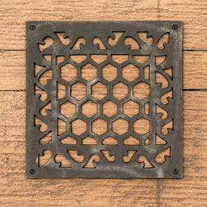 Puede incluir: Una rejilla de ventilación cuadrada de metal gris oscuro con un patrón central de panal rodeado por un borde decorativo de volutas. La rejilla está asegurada en cada una de sus cuatro esquinas con pequeños tornillos oscuros de cabeza plana.