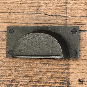 Puede incluir: Un tirador de cazoleta rectangular de metal gris oscuro con una zona de agarre semicircular y una placa de montaje plana. El herraje incluye cuatro orificios para tornillos visibles, uno en cada esquina, con tornillos de cabeza plana instalados.