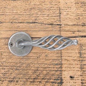 Könnte beinhalten: Ein silberfarbener Türgriff aus Metall mit einer runden Montageplatte, einer sichtbaren Schraube und einem horizontalen Hebel, der ein dekoratives, offenes, gedrehtes Käfigdesign aufweist und zu einem abgerundeten Ende hin schmaler wird.